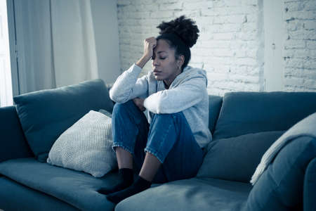 Depressed african woman with sad expression, headache and depression feeling desperate at home. Afro American teen or Attractive black young female lying thoughtful and sick on sofa in pain and worry.の写真素材