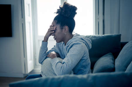Depressed African woman with sad expression, headache and depression feeling desperate at home. Afro American, Attractive black young female lying thoughtful and sick on sofa in pain and worry.の写真素材