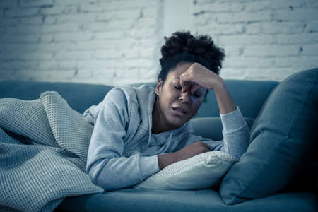 Depressed African woman with sad expression, headache and depression feeling desperate at home. Afro American, Attractive black young female lying thoughtful and sick on sofa in pain and worry.の写真素材