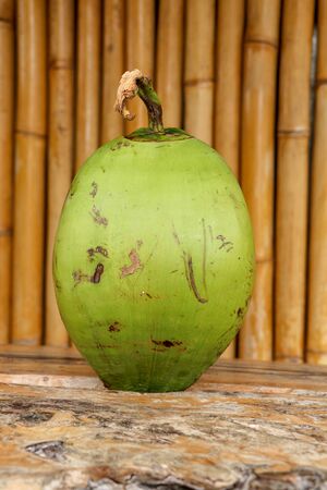 Fresh young coconut on table with massive wooden board. Young Coconut Fruit for Drink Sweet Juice and Eat. Healthy natural refreshment full of energy and vitamins. In the background wall of bamboo.の写真素材