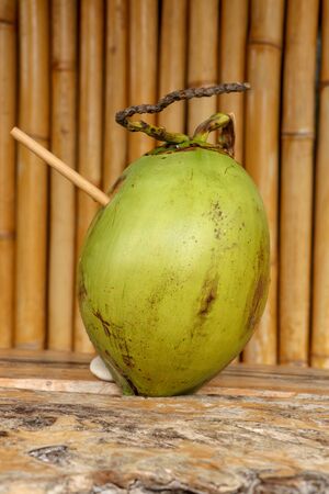 Fresh young coconut with bamboo straw on table with massive wooden board. Healthy natural refreshment full of energy and vitamins. In the background wall of bamboo trunks. Nice tropical concept.の写真素材