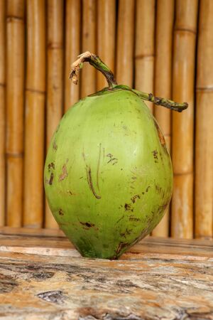 Fresh young coconut on table with massive wooden board. Young Coconut Fruit for Drink Sweet Juice and Eat. Healthy natural refreshment full of energy and vitamins. In the background wall of bamboo.の写真素材