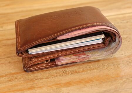 Brown leather wallet full of banknotes and credit cards. Men's wallet on a wooden table. Money in a closed brown purse. Best background for your project.の写真素材