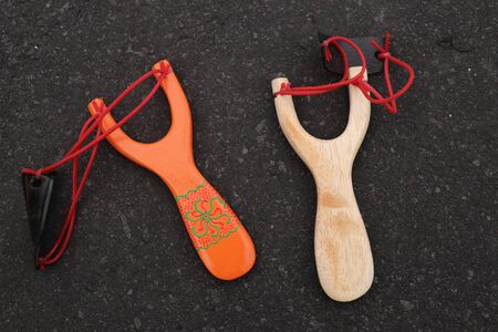 Couple of simple wooden slingshot isolated. Orange and natural wooden handle with two red rubber bands attached to a black leather strip used to put things to be shot on black background. Primitive but effective weapon.の写真素材