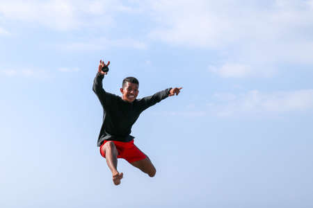 Man jumping happy on the beach with a blue sky in the background. Young cheerful Indonesian jumps. A teenager in a good mood jumps in the air. Background with blue sky. Sunny tropical day.の写真素材