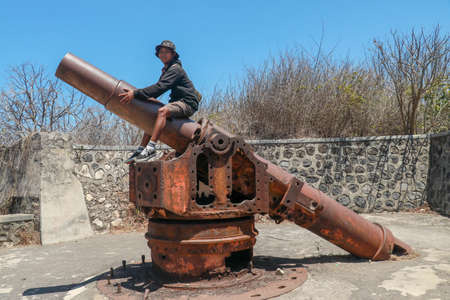 An Asian teenager sits on a historic World War II cannon. Military stone fortifications from World War II. A young guy sitting on an ancient cannon. Lombok, Indonesiaの写真素材