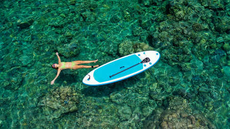 Aerial view of pretty woman lying on azure sea surface. Calm Relaxed Woman Resting in calm sea by Sup board. Funny summer water sports. Healthy Lifestyle. Surfing, Fashion. Summer Vacation. 4Kの写真素材
