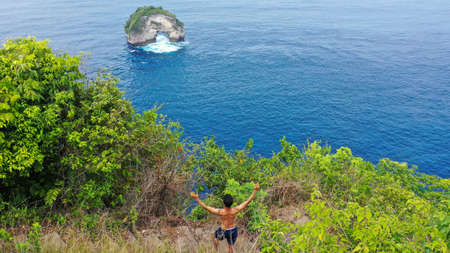 A young man stands on a cliff above the ocean and raises his hands. Tourist stands on a hill with stunning sea views. Travel, adventure, tourism conceptの写真素材