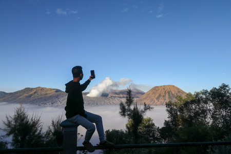 Young asian man taking a picture of beautiful mountains landscape in Bromo Mountain National Park, East Java, Indonesiaの写真素材