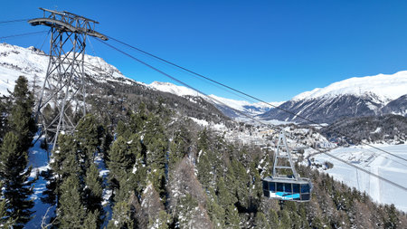 Cable car in the mountains in the winter forest. The cabins move along the gondola road through the winter snow forest. White clouds covered mountains. Top view from a drone. St. Moritzの写真素材