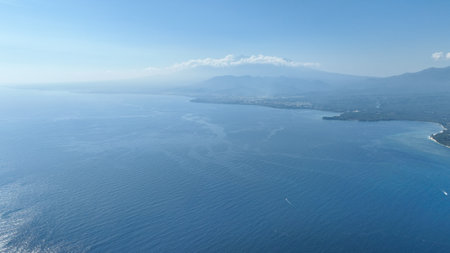 A breathtaking aerial view of the coastline of Lombok island, where the deep blue sea meets lush green land. The distant mountains shrouded in mist add a sense of mysteryの写真素材