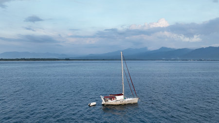 Aerial view of sailboat moored on calm blue water. Cloudy sky forming a breathtaking backdrop. Small motor boat parked in the sea opposite the coast of a tropical islandの写真素材