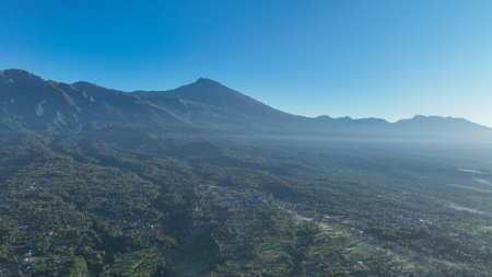 The majestic silhouette of Mount Rinjani volcano rises above a sea of green forest, with misty...