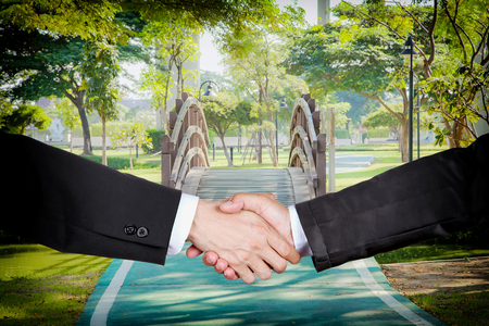 Businessmen shaking hands with a small curved wooden bridge in the garden background.の写真素材