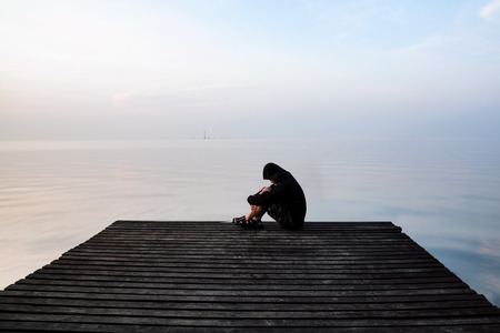 Frustrated depression man in the hood sitting on wooden bridge, near the beach on sunset. Concept of unemployed, sadness, depressed and human problemsの写真素材