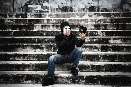 Asian man in the black hood and white mask with confused depression sitting on stairs and looking at black mask, in scary abandoned building, Human face expression, Good and Bad temptation concept.の写真素材
