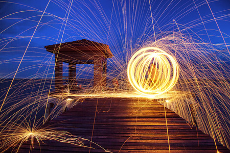 Hot Golden Sparks Flying from Man Spinning Burning Steel Wool on Wooden Bridge Extended into the Sea., Long Exposure Photography using Steel Wool Burning.の写真素材