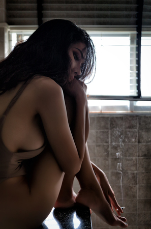 Beautiful lonely woman in frustrated depression sitting on wash basin counter in the bathroom, Hand holding smoke cigarette, Concept of unemployed, sadness, depressed and human problems in dark tone.の写真素材
