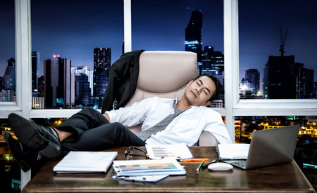 Feeling exhausted.,Young Asian businessman sitting on couch in office., Sleeping while working with computer at his working place., On the desk overwhelmed by paperwork.の写真素材
