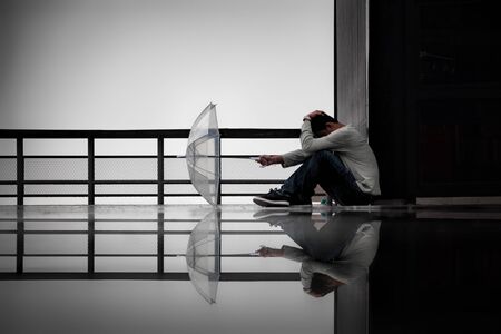Depressed man with transparent umbrella in hand put on the ground., Sitting alone on wet floor with water reflection in rainy., Concept of Major depressive disorder, sadness, depression and problems.の写真素材