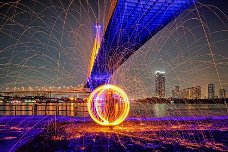 Hot Golden Sparks Flying from Man Spinning Burning Steel Wool under Bhumibol Bridge in Bangkok Thailand., Long Exposure Photography using Steel Wool Burning.の写真素材
