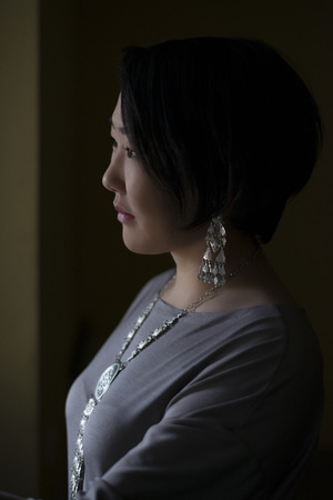 Asian girl looking out the window standing in a dark room.の写真素材