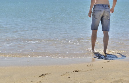 Young man in pair of shorts seen of back walking at the water s edgeの写真素材