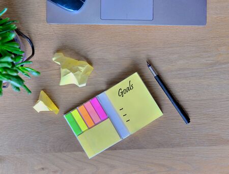 yellow note paper and pen with other crumpled paper on a wooden desk with plant and computer 