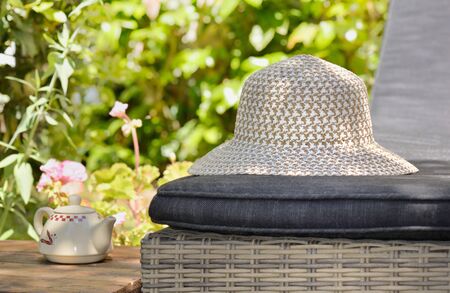 close on braided hat put on a lounge chair next to wooden table with teapot in gardenの写真素材