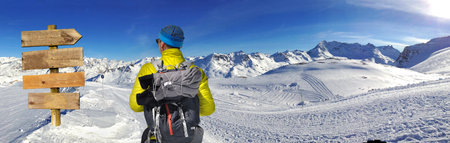 view on back of a hiker looking at a wooden panel directional wooden and panoramic view on snowy peak mountain under sunny blue skyの写真素材