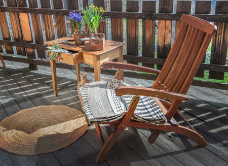 wooden deck chair next to a table with flowers in a pot on a terrace in front of fenceの写真素材