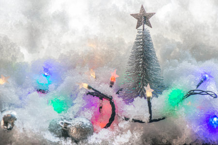 Christmas decoration in the snow with litle balls and silver christmas tree with colorful light of garland in front of a glass of windowの写真素材