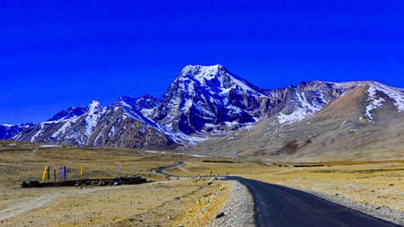 A long lonely road with curves on the tibetan plateau with snow clad mountains and peaks and clear blue sky in front and tibetan prayer flags on one sideの写真素材