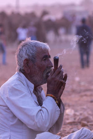 High dynamic range image of a rural Indian man siting an smoking tobacco inside a pipe at Pushkar, Rajasthan, India on 19 November 2018のeditorial素材