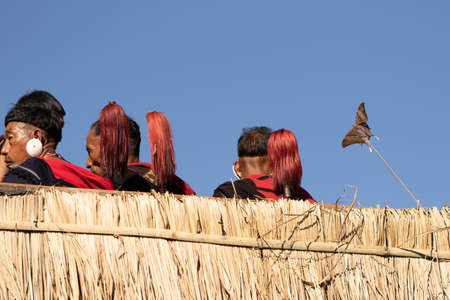 Naga men siting with only their heads  and their traditional weapons visible behind thatched fence and a butterfly stuck in a twig at a village at kohima Nagaland India on 4 December 2016のeditorial素材
