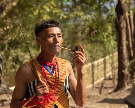 A Naga Tribesman dressed in tribal Attire holding a tribal weapon in one hand and smoking a pipe at Kisama village in Nagaland India on 2 December 2016のeditorial素材