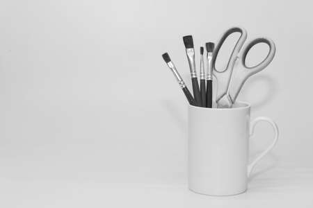 Paint brushes and a scissors kept inside a white ceramic mug in monochrome with copy spaceの写真素材