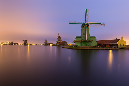 Night time landscape of the river Zaan with four windmills lined up at 'De Zaanse Schans' in Zaandam, the Netherlands. Lights from street lamps and houses are reflected on the water surface. Sky is brightly colored in a gradient by city lights combined wiの写真素材