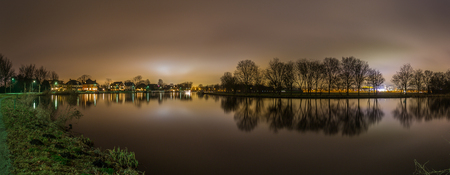 Peaceful panoramic view of the Amstel river and trees while city lights from Amsterdam illuminate the night sky in the backgroundの写真素材