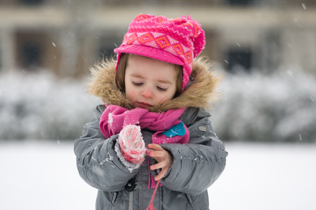 Portrait of a young infant outside in snowy weather dressed in winter coat, scarf and cap. The girl is looking captivated at snowflakes falling on her glovesの写真素材