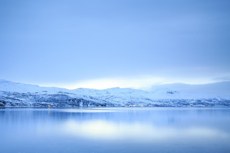 Typical norwegian coastline with rocks at low tide at a fjord in Troms county, Norway. The sun is low above the horizon, the sky clouded. Mountains are covered with snow during the cold winter.の写真素材