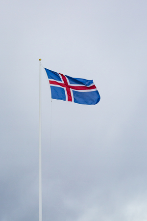 A civil flag and ensign Iceland against a clouded sky. The civil national flag of Icelanders is blue as the sky with a snow-white cross, and a fiery-red cross inside the white cross.の写真素材