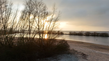 Tranquil sunset over a calm lake with silhouettes of bare trees and a sandy shore in the foreground, depicting a serene natural landscape.の写真素材