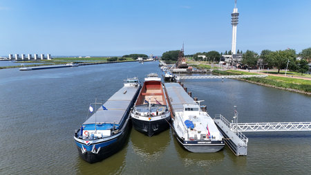 Three cargo ships docked at a river port with a tall communication tower in the background.の写真素材