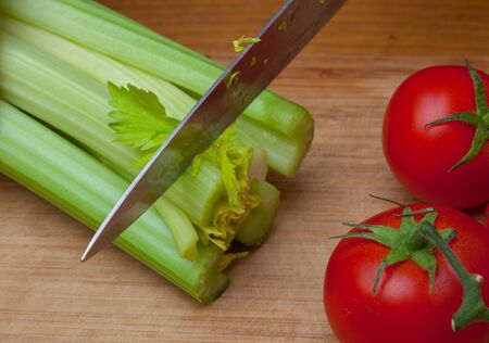 Green stalks of celery and red tomatoes on a wooden cutting board beside an iron knifeの写真素材