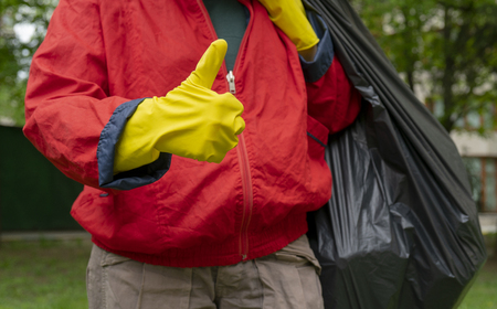 Garbage collection. Man in red coat and yellow gloves showing thumb up, carries a big black bag with trash.の写真素材