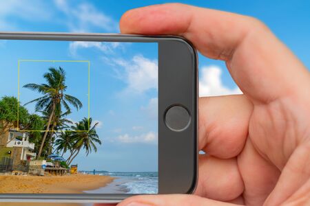 Beautiful coconut tree on background of blue tropical sky. Ocean waves on beach. Smartphone in hand....
