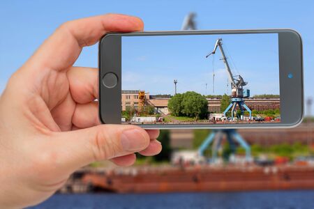Cranes operate in river port. Cargo port on river. Construction crane in screen of smartphone.の写真素材