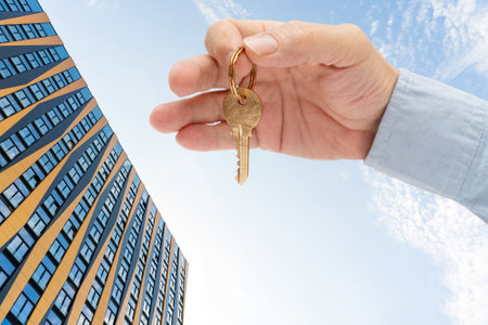 Apartment key in a man's hand. Brass house door lock key. Modern building against the blue sky. View from below.の写真素材