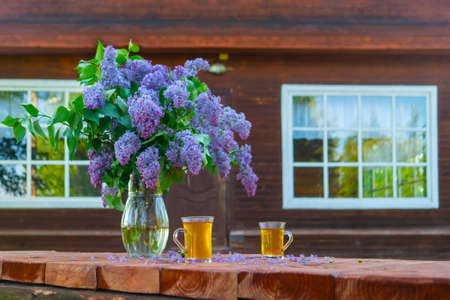 A bouquet of beautiful lilacs in a glass vase stands on a wooden table. Wooden cottage in the forest.の写真素材